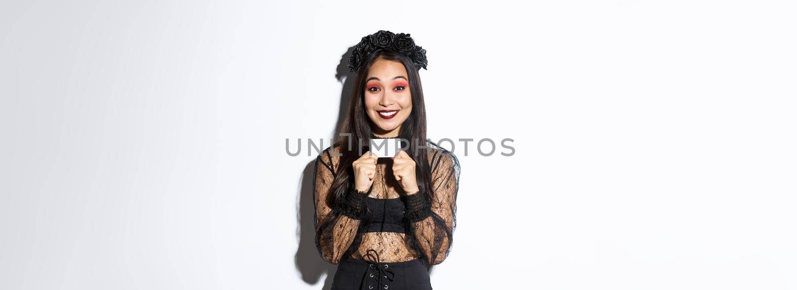 Excited smiling asian girl in black wreath and gothic witch costume showing credit card, standing over white background.