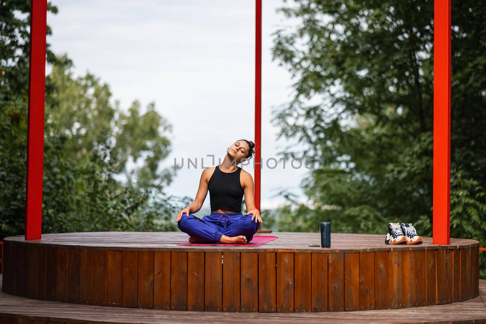Young woman is practising yoga sitting in the lotus position on a wooden podium in the garden