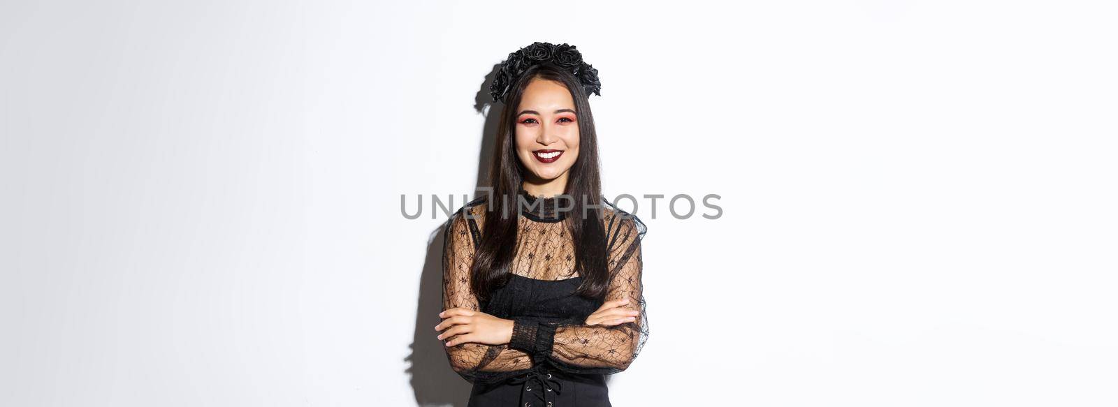 Smiling beautiful asian woman celebrating halloween, wearing black wreath and dress, gothic makeup, looking happy at camera. Female celebrating autumn event, trick or treating in costume by Benzoix