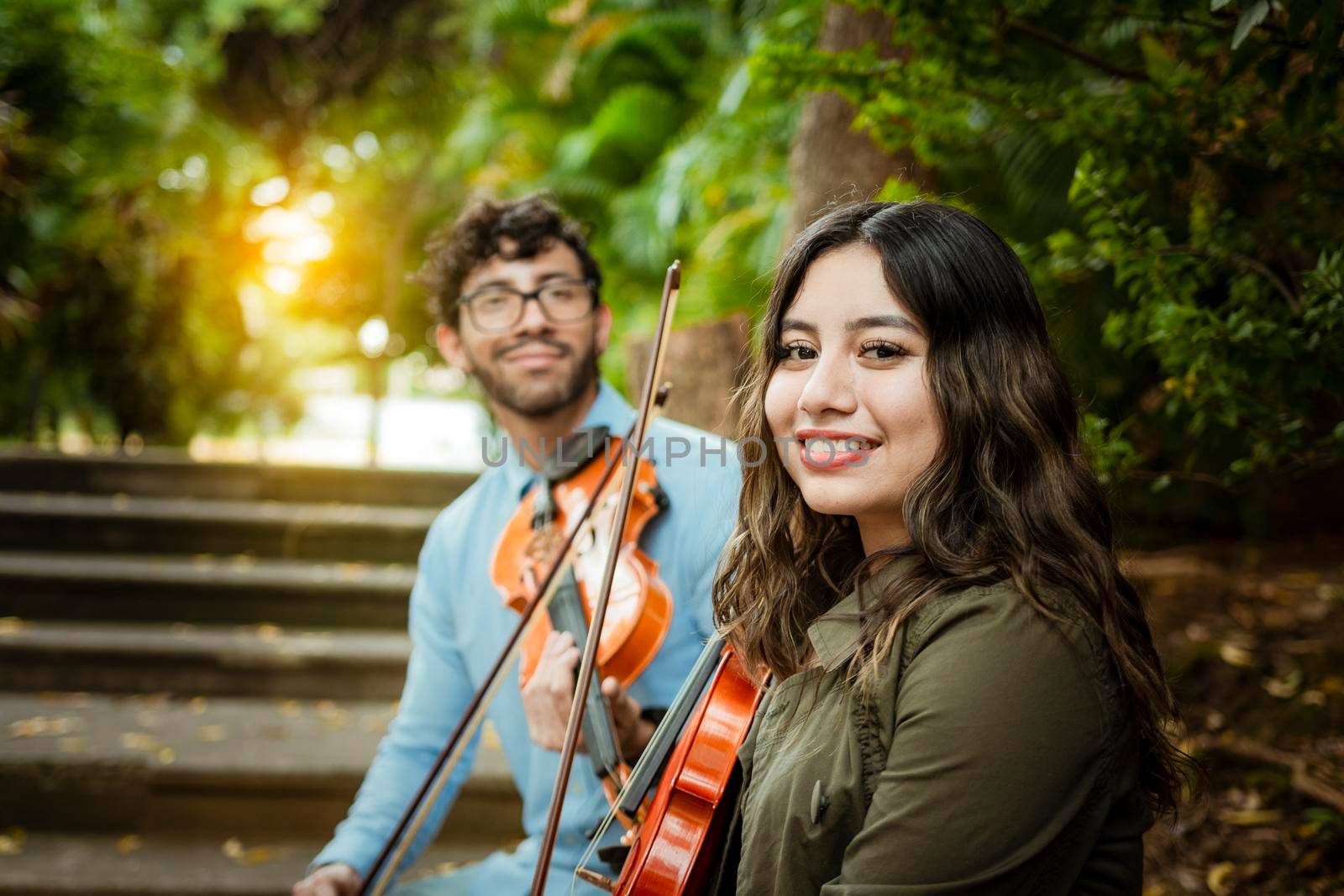 Two young violinists sitting outdoors looking at the camera. Portrait of two young violists sitting outside. Man and woman violinist sitting on the stairs. Concept of two young violinists by isaiphoto