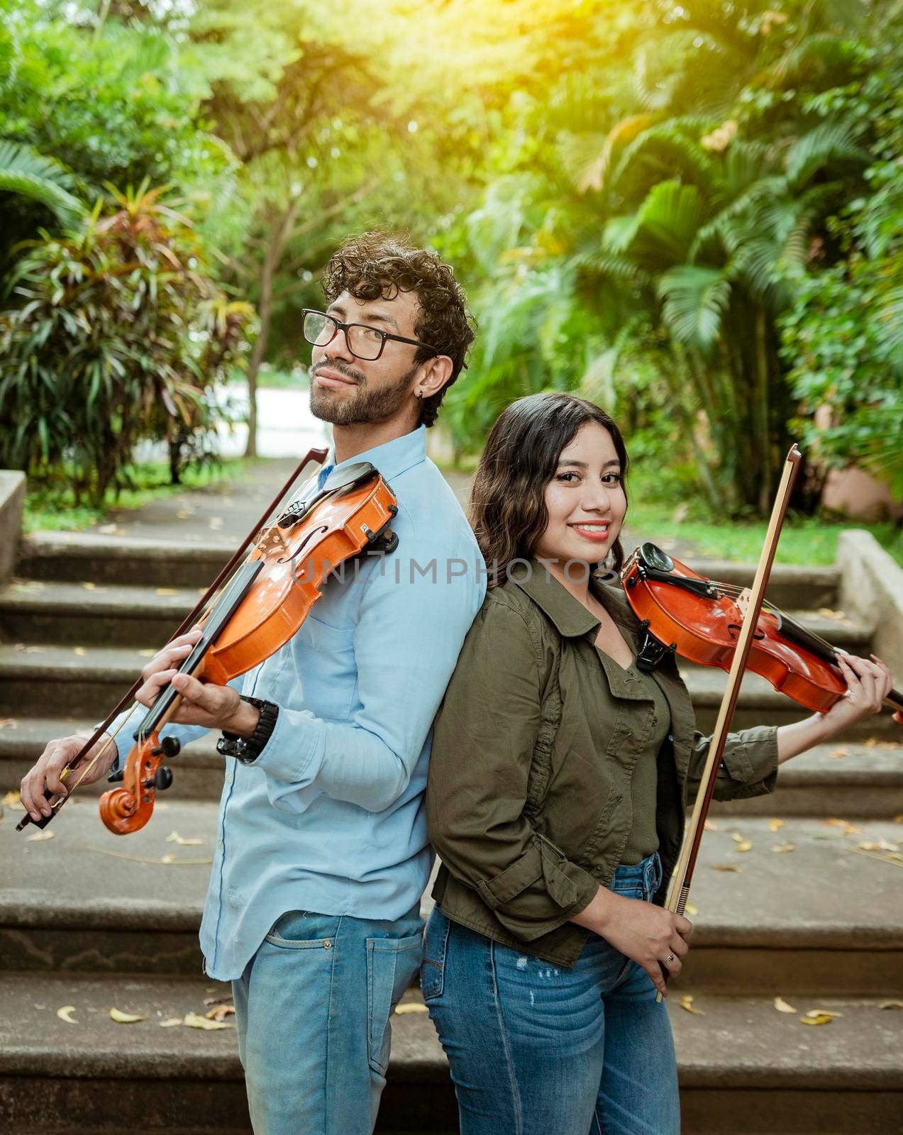 Portrait of two young violists back to back outdoors. Portrait of smiling male and female violinist back to back outdoors. Two young violinists back to back holding their violins by isaiphoto