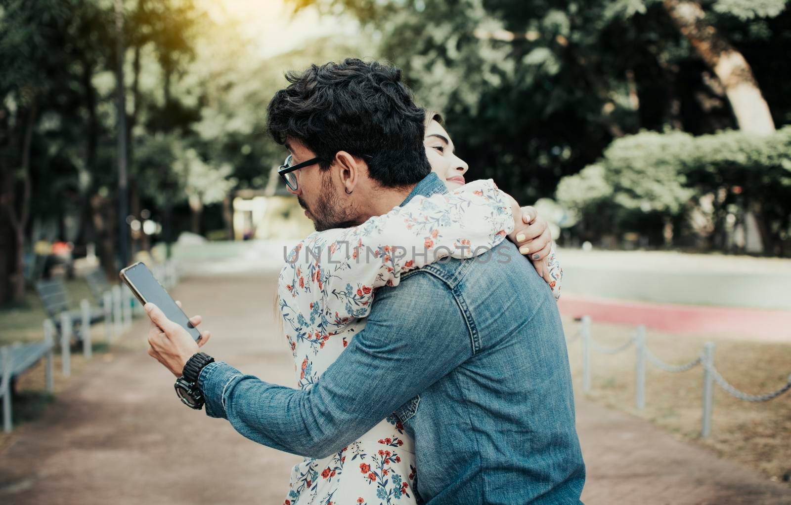 Unfaithful man hugging his girlfriend and looking at the cell phone. Concept of unfaithful man using cell phone. Unfaithful man looking at the cell phone while hugging his girlfriend outdoors