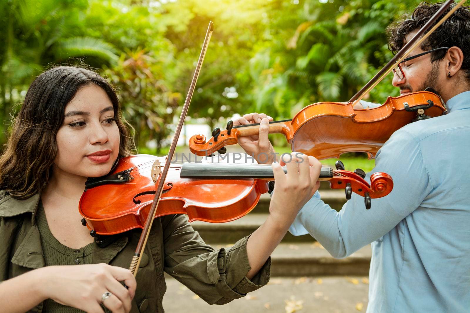 Violinist man and woman back to back playing violin in a park outdoors. Two young violinists standing playing violin in a park. Portrait of man and woman together playing violin in park