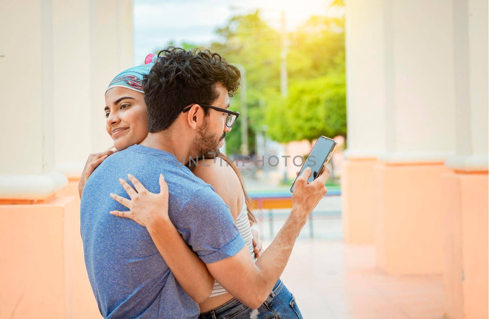 Unfaithful boyfriend hugging his girlfriend and looking at the cell phone. Concept of unfaithful man using cell phone. Unfaithful man looking at the cell phone while hugging his girlfriend outdoors