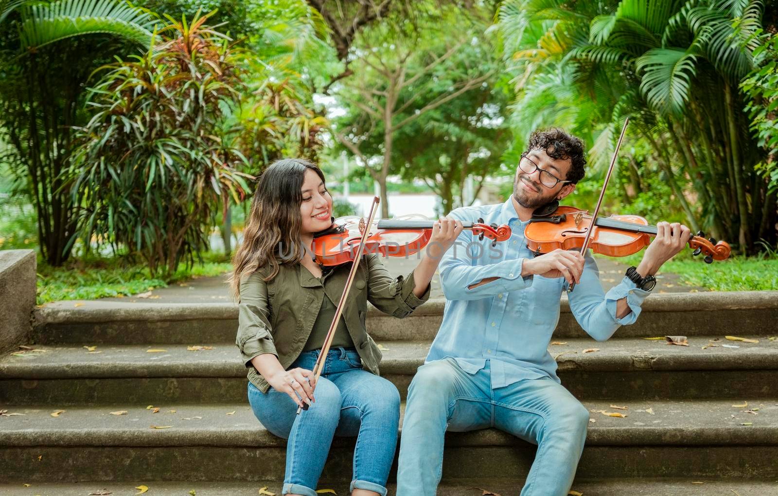 Portrait of two young violists playing violin sitting on stairs outside. Portrait of smiling violinists sitting on stairs playing violin. Man and woman sitting on stairs playing violin by isaiphoto