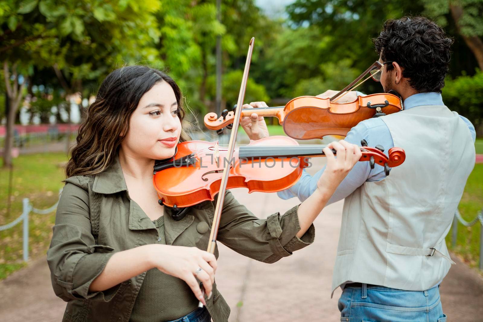 Two young violinists standing playing violin in a park. Portrait of man and woman together playing violin in park. Violinist man and woman back to back playing violin in a park outdoors by isaiphoto