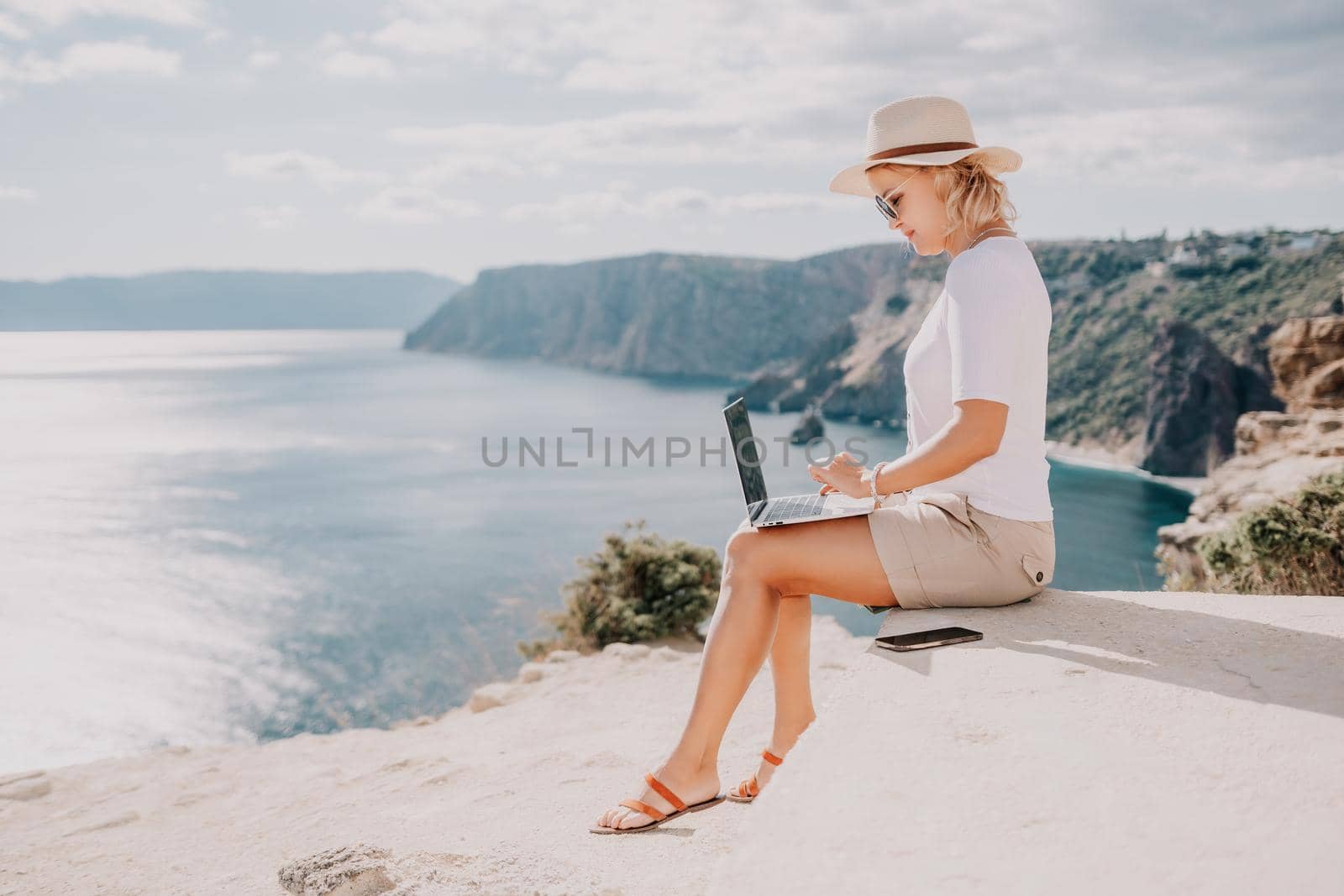 Successful business woman in yellow hat working on laptop by the sea. Pretty lady typing on computer at summer day outdoors. Freelance, travel and holidays concept.