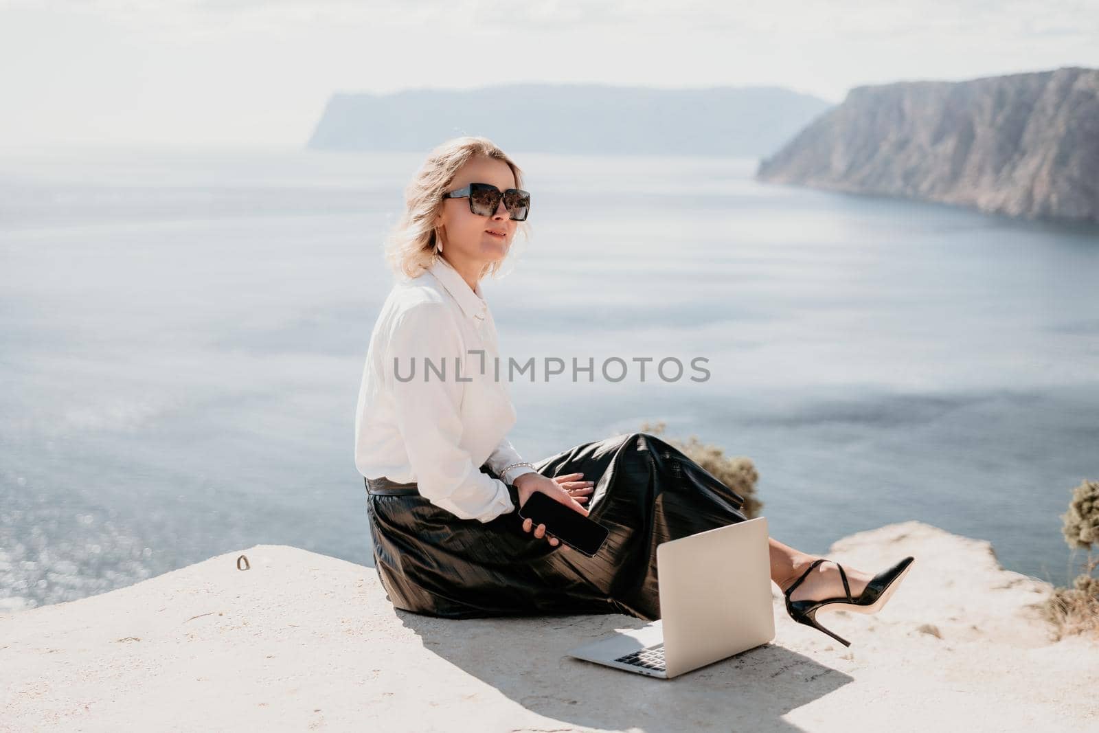 Successful business woman in yellow hat working on laptop by the sea. Pretty lady typing on computer at summer day outdoors. Freelance, travel and holidays concept.