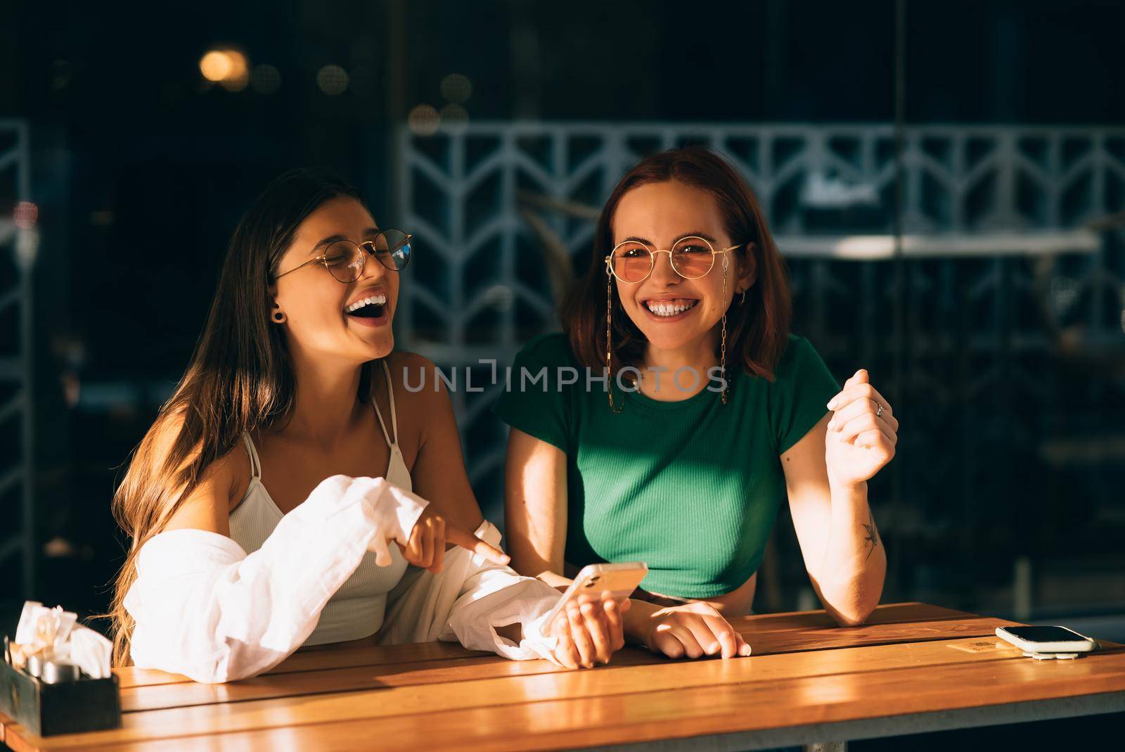 Two excited young girls using mobile phones while sitting at the cafe outdoors