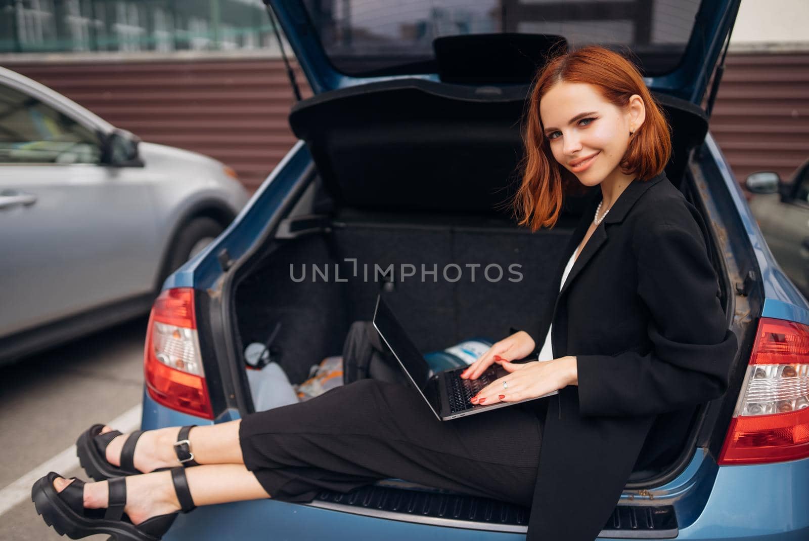 Business attractive young woman working on laptop while sitting in trunk of car while traveling