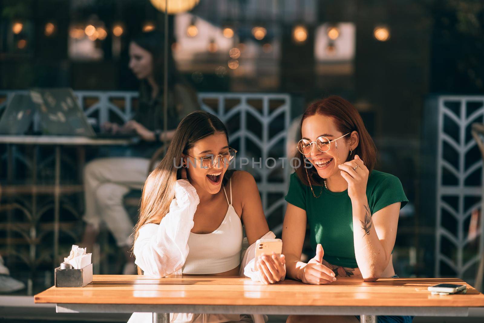 Two excited young girls using mobile phones while sitting at the cafe outdoors