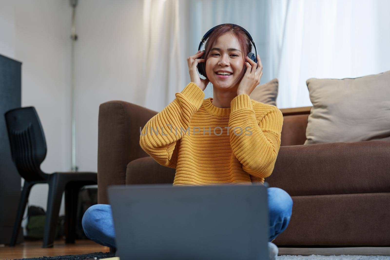 A portrait of a young Asian woman with a smiling face wearing a pair of headphones and using computer and listening to music while sitting on the sofa.