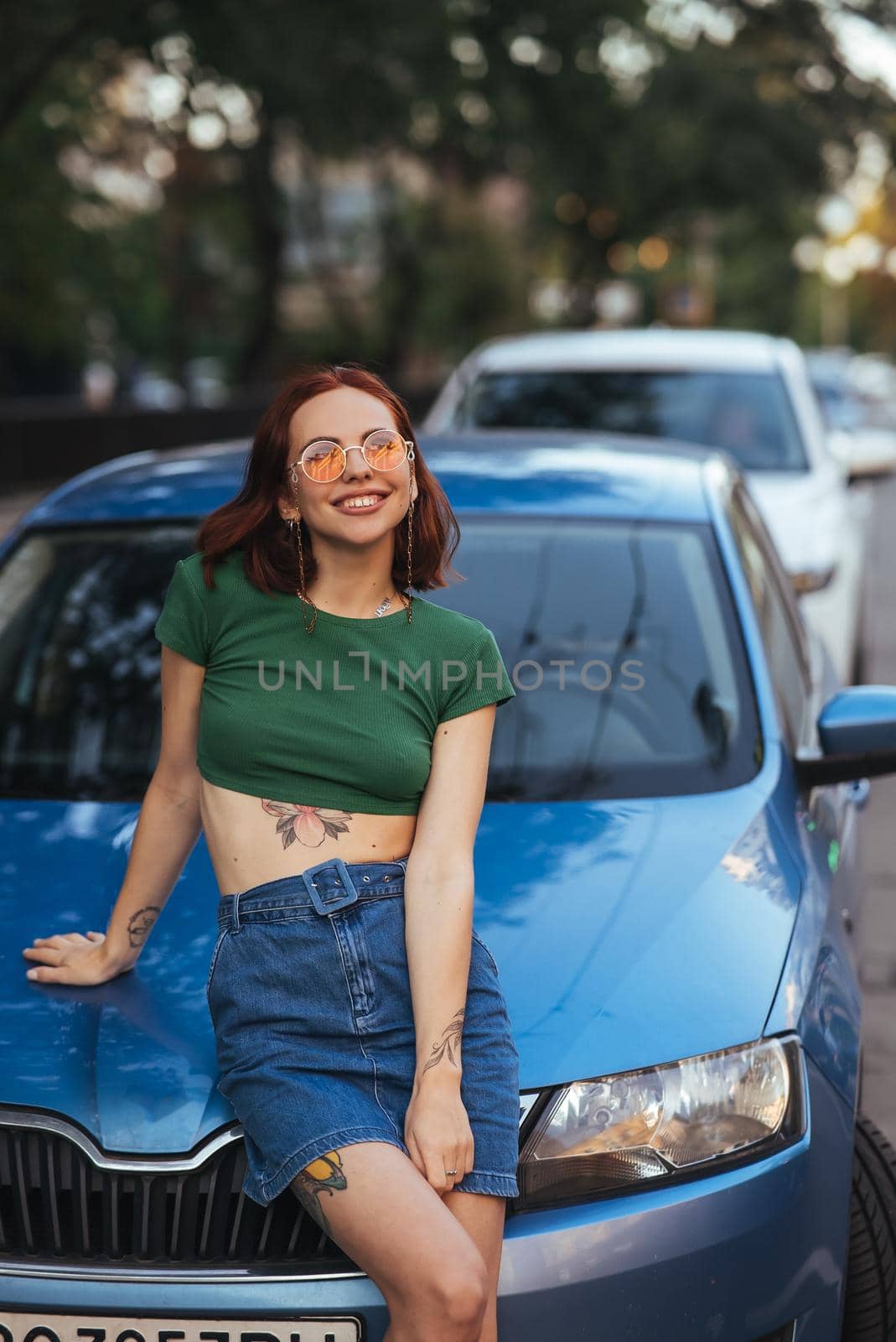 Beautiful girl in sunglasses sits on the hood of a blue car, against the road
