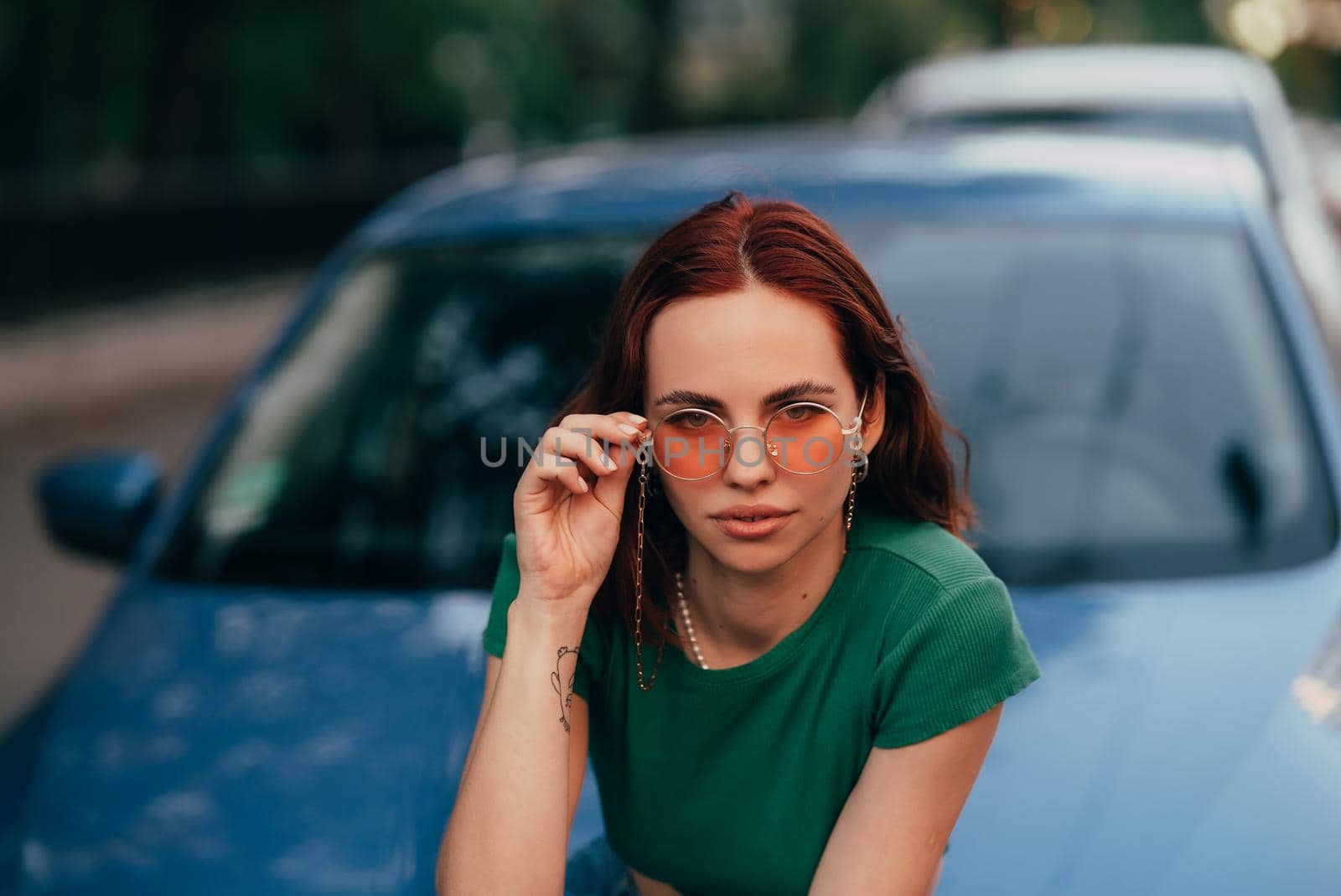 Beautiful girl in sunglasses sits on the hood of a blue car, against the road