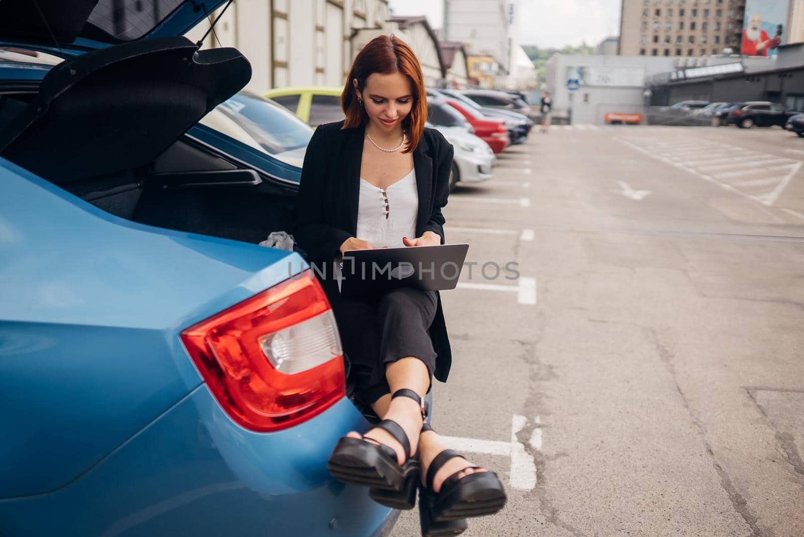 Business attractive young woman working on laptop while sitting in trunk of car while traveling