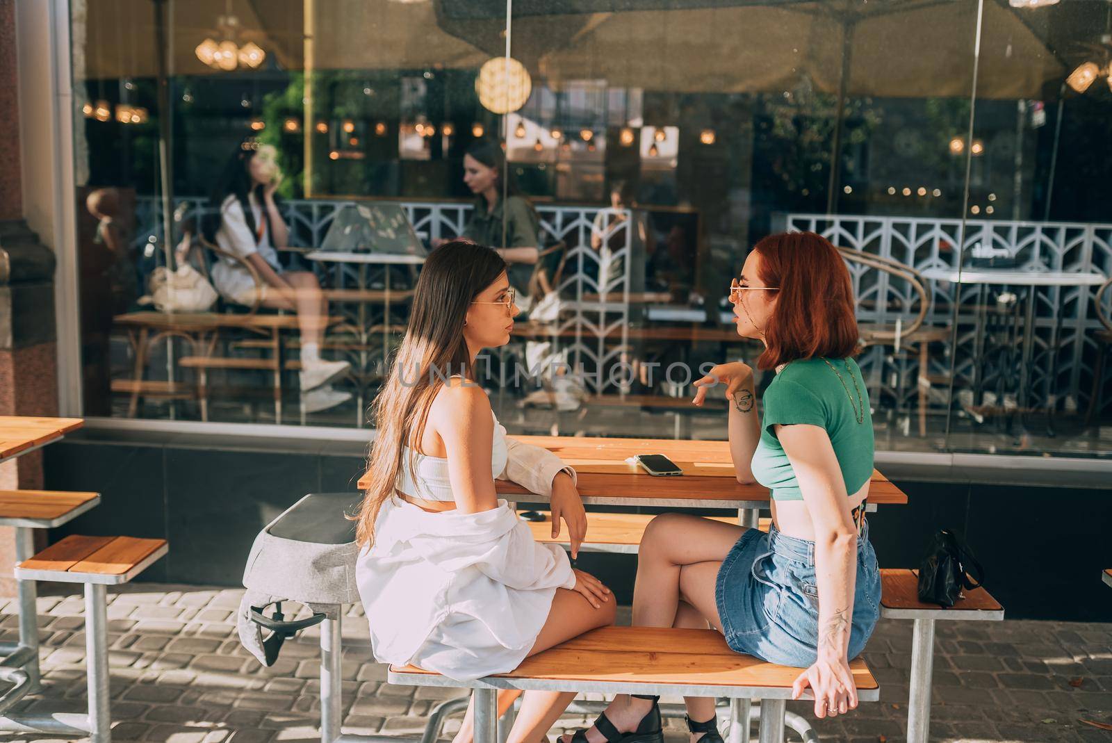 Two pretty girlfriends talking while sitting in a bar at the city street