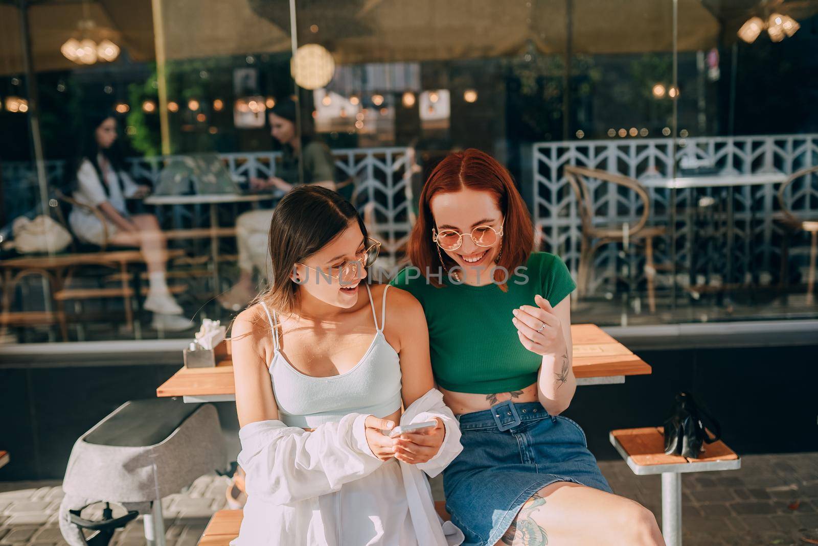 Two excited young girls using mobile phones while sitting at the cafe outdoors
