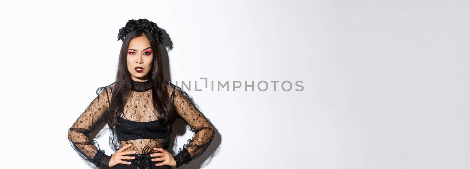 Angry and pissed-off asian female magician, evil witch in black dress and wreath looking mad at someone, frowning and looking judgemental, standing over white background by Benzoix