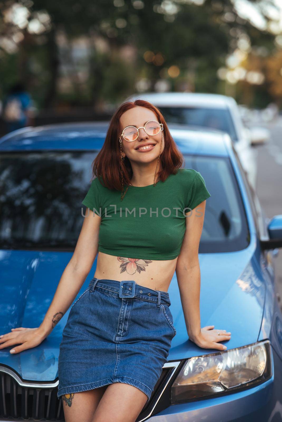 Beautiful girl in sunglasses sits on the hood of a blue car, against the road