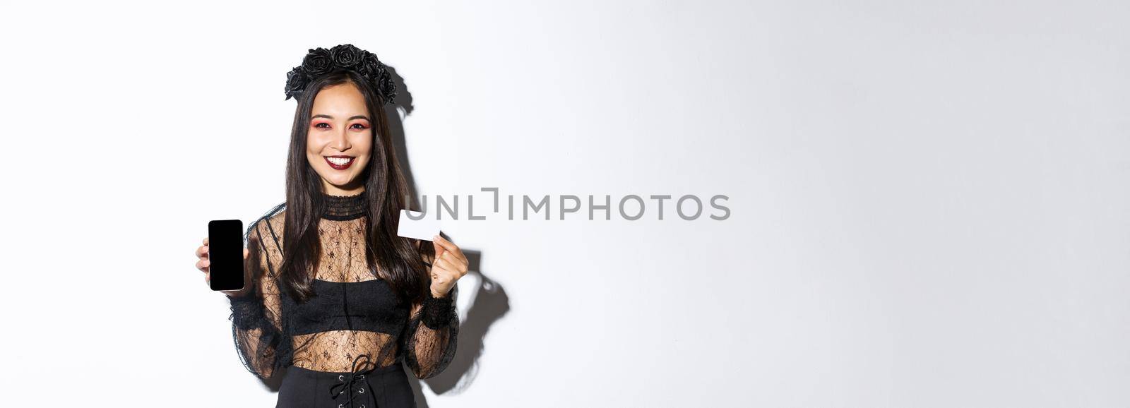 Smiling attractive woman in black gothic dress showing credit card and smartphone display, standing over white background in halloween costume by Benzoix