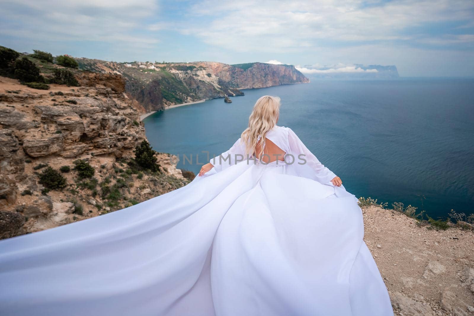 Blonde with long hair on a sunny seashore in a white flowing dress, rear view, silk fabric waving in the wind. Against the backdrop of the blue sky and mountains on the seashore