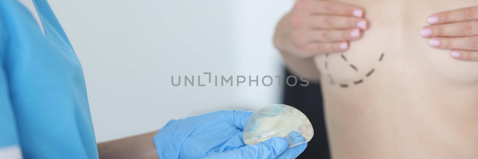 Close-up of doctor hold silicone breast implant and client with marking on breast. Woman topless in cabinet. Surgery, plastic operation, health concept