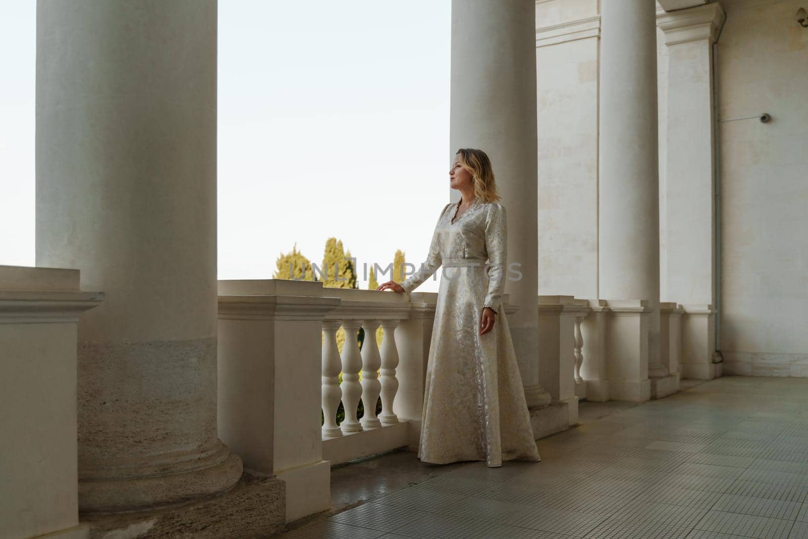 a beautiful mature woman in a long white dress stands on the balcony and looks into the distance.