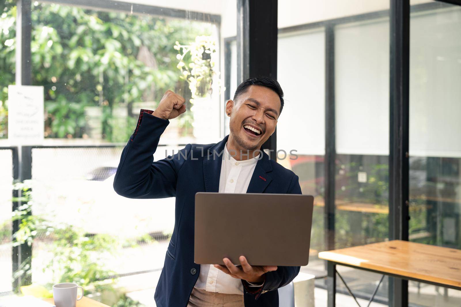 Happy young businessman in suit looking at laptop excited by good news online, lucky successful winner man standing at office desk raising hand in yes gesture celebrating business success win result.