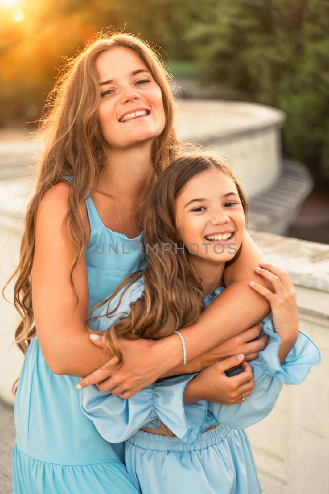 Portrait of mother and daughter in blue dresses with flowing long hair against the backdrop of sunset. The woman hugs and presses the girl to her. They are looking at the camera