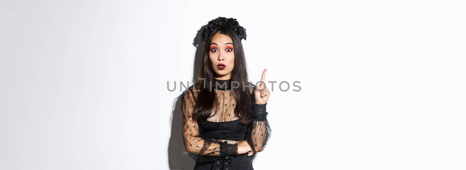 Beautiful young asian woman in black lace dress and gothic makeup have thought. Girl in witch costume raising finger, suggesting halloween idea.