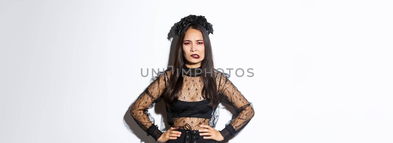 Annoyed and bothered evil witch looking complicated and biting lower lip, standing over white background in black lace dress and wreath. Girl in halloween costume trick or treating.