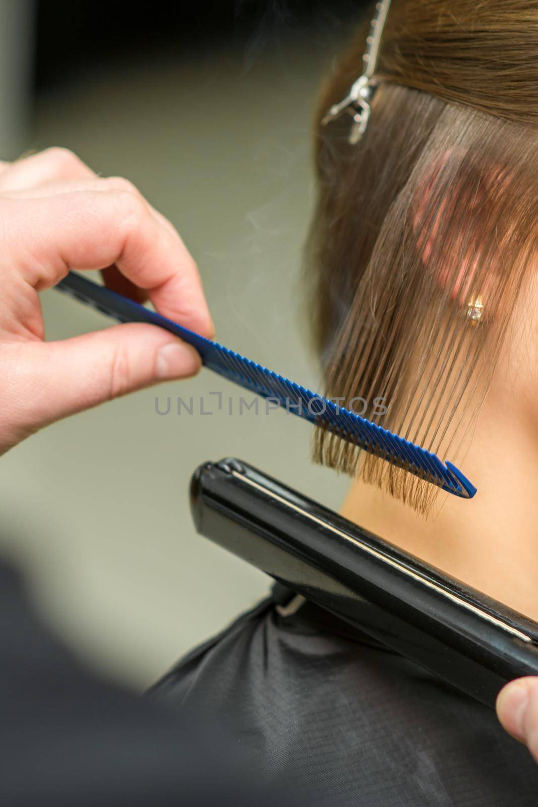 Hairstylist is straightening short hair of young brunette woman with a flat iron in a hairdresser salon, close up. by okskukuruza