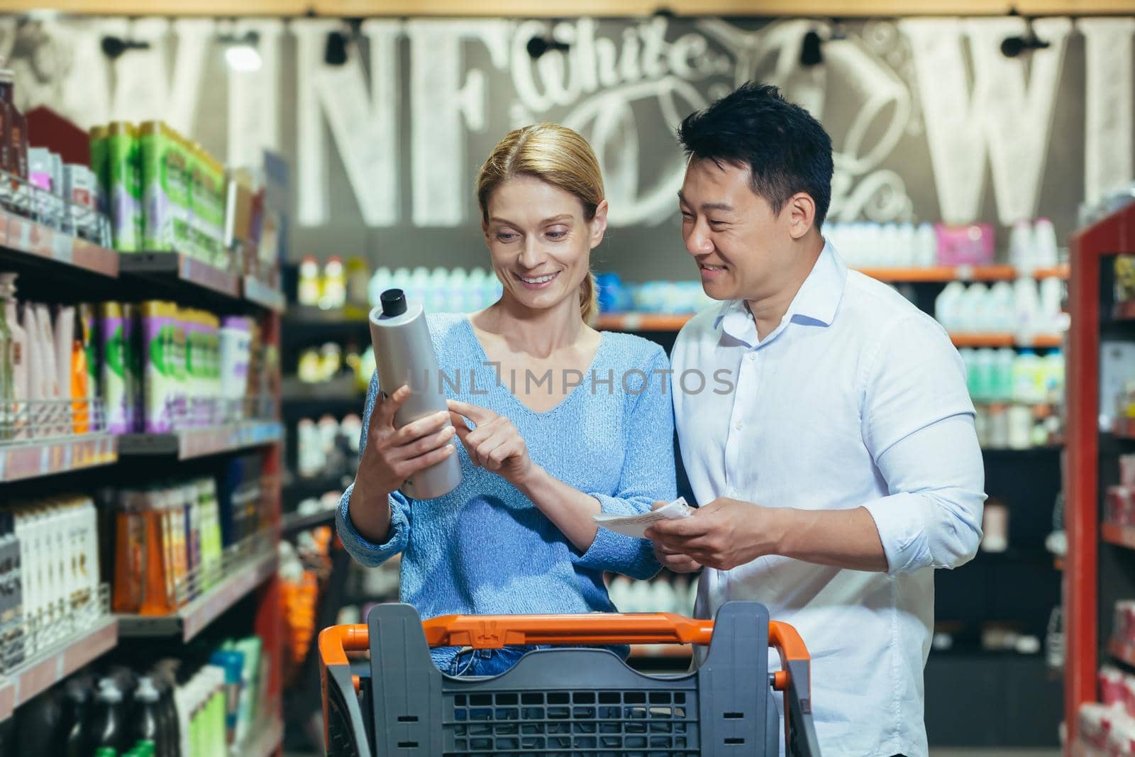 A young interracical couple, a woman and an Asian man walk between the rows with a trolley in a supermarket, the woman holds a bottle of shampoo in her hands, reads the composition on the label.