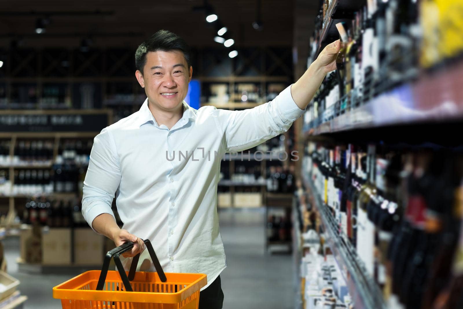 A young handsome Asian man businessman in a white shirt chooses wine in the alcohol section of a supermarket, takes one bottle. He holds a product basket in his hand, looks at the camera, and smiles.