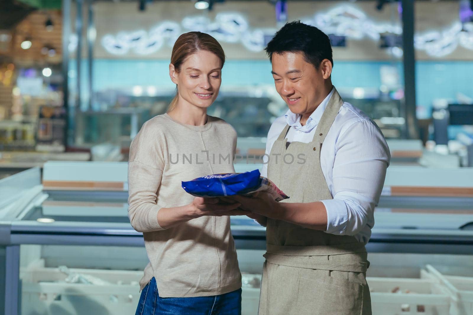 Consultant in a supermarket. A friendly Asian man in an apron offers a young beautiful woman to buy a blue pack of frozen food in the store.