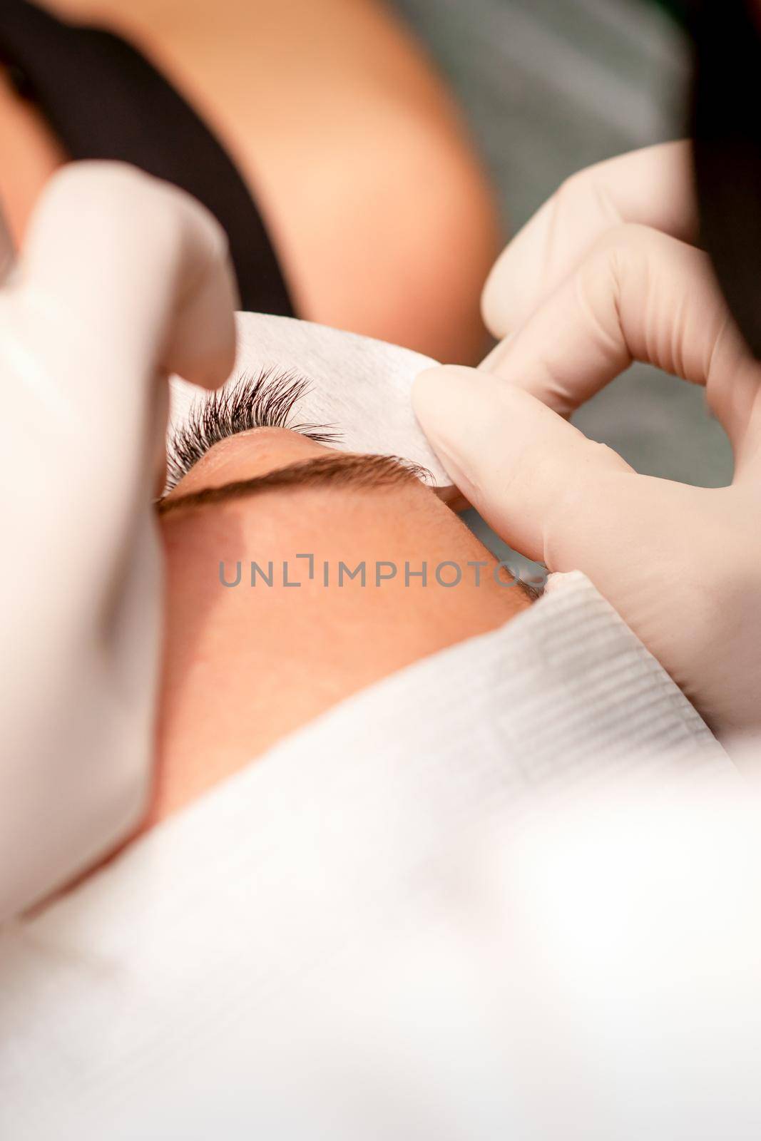 The hands of the cosmetologist are gluing white tape under the eye of the young caucasian woman during the eyelash extension procedure, closeup. by okskukuruza