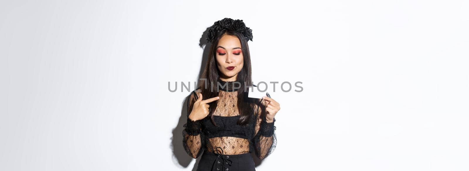 Image of curious beautiful asian girl in witch costume pointing finger at credit card, standing over white background.
