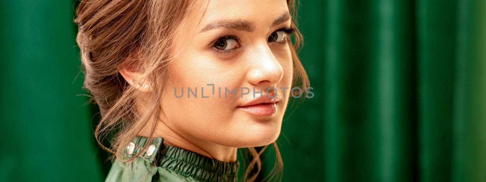 The fashionable young woman. Portrait of the beautiful female model with long hair and makeup with closed eyes. Beauty young woman with a brown curly hairstyle on the background of a green curtain