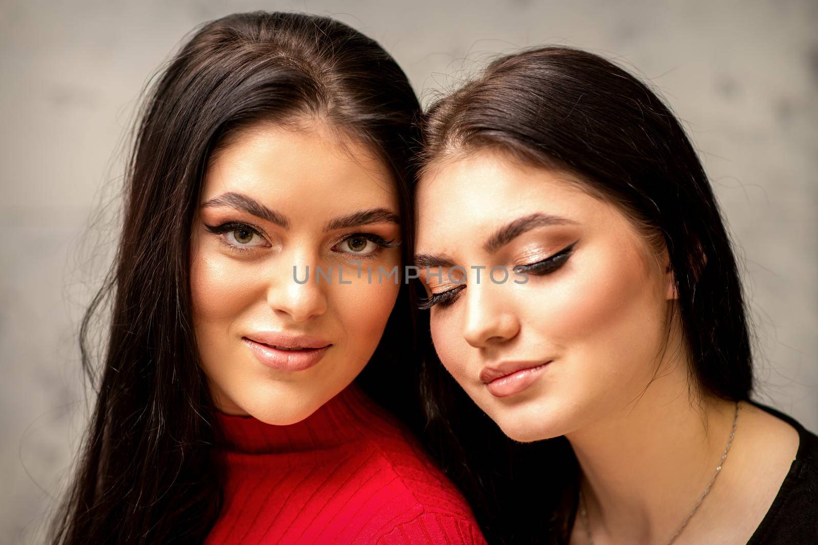 Two fashionable young women. Portrait of the two beautiful female models with long hair and makeup. Two beauty young caucasian women with a black hairstyle on the background of a gray wall