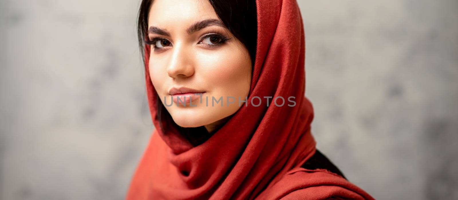 The fashionable young woman. Portrait of the beautiful female model with long hair and makeup in a red scarf. Beauty young caucasian woman on the background of a gray wall. by okskukuruza