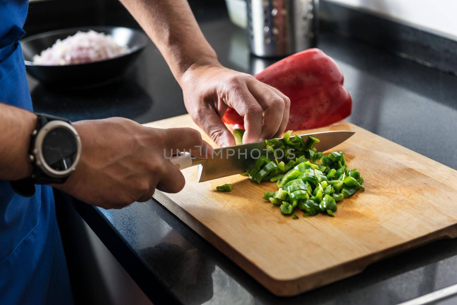 close-up of male hands chopping green and red pepper on a wooden board with a chef's knife, he has other vegetables needed for his recipe in the background