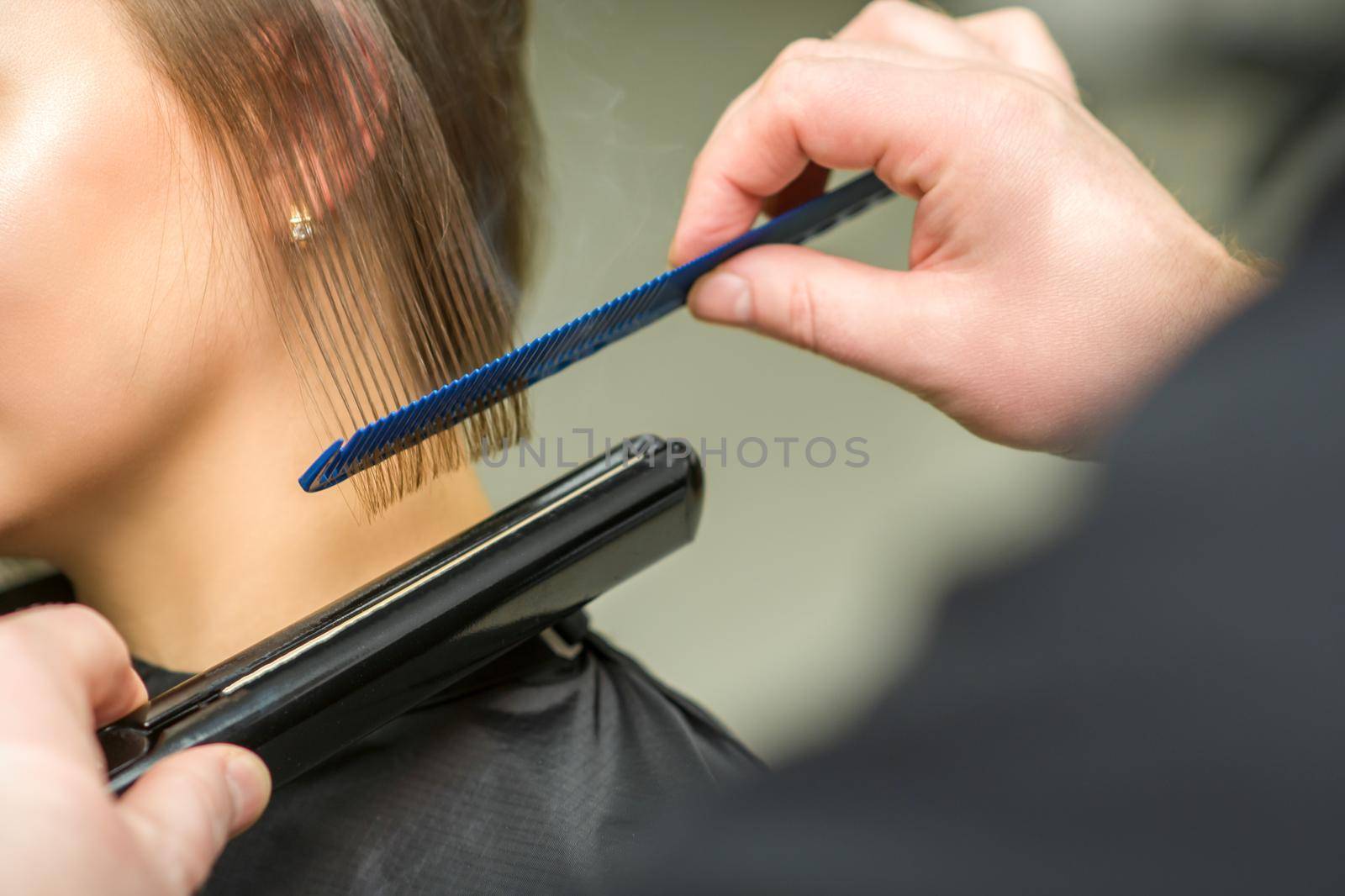 Hairstylist is straightening short hair of young brunette woman with a flat iron in a hairdresser salon, close up. by okskukuruza