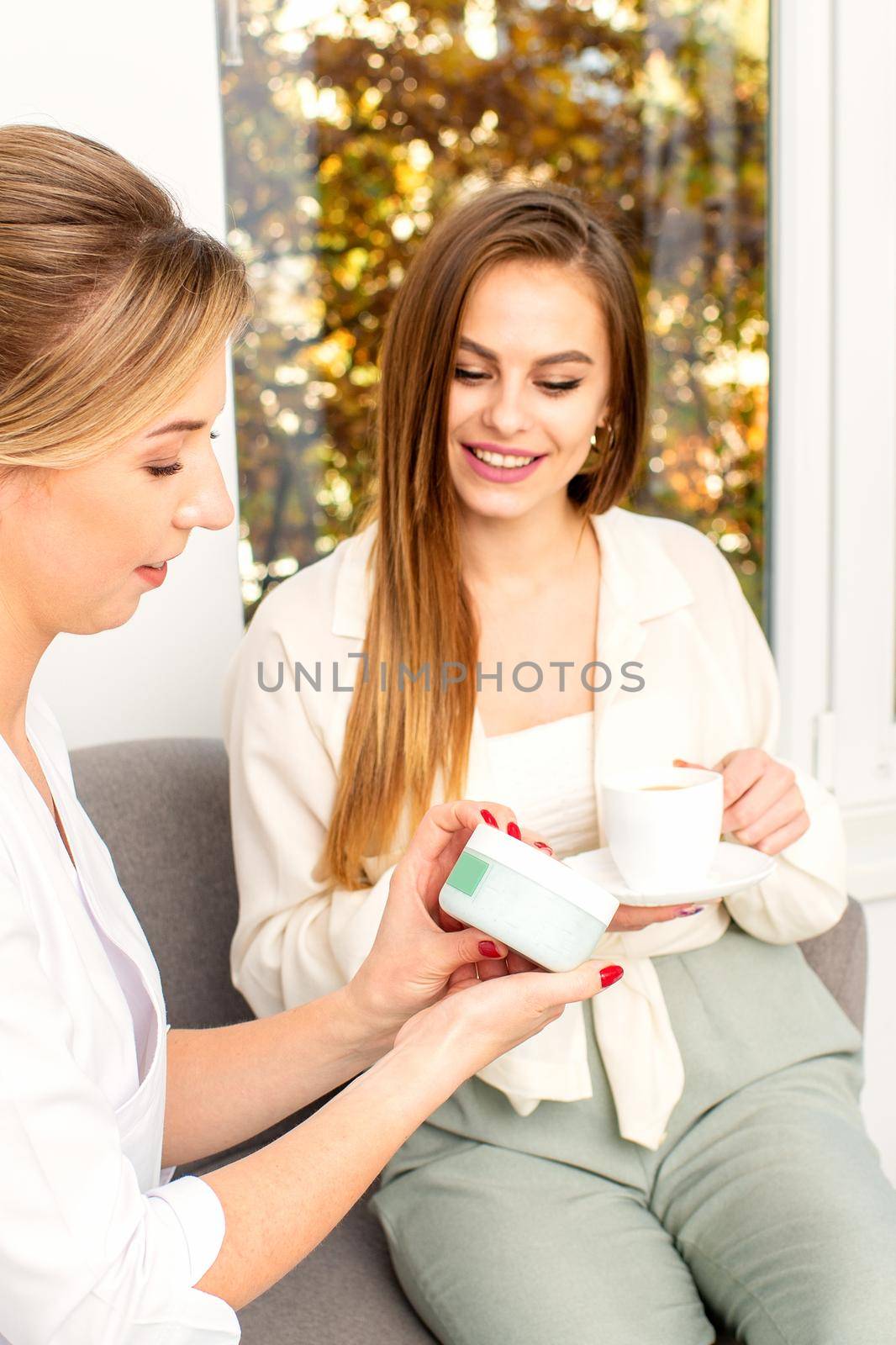 Beautician offering product for the young woman holding a white plastic jar with a cream sitting on the sofa. by okskukuruza