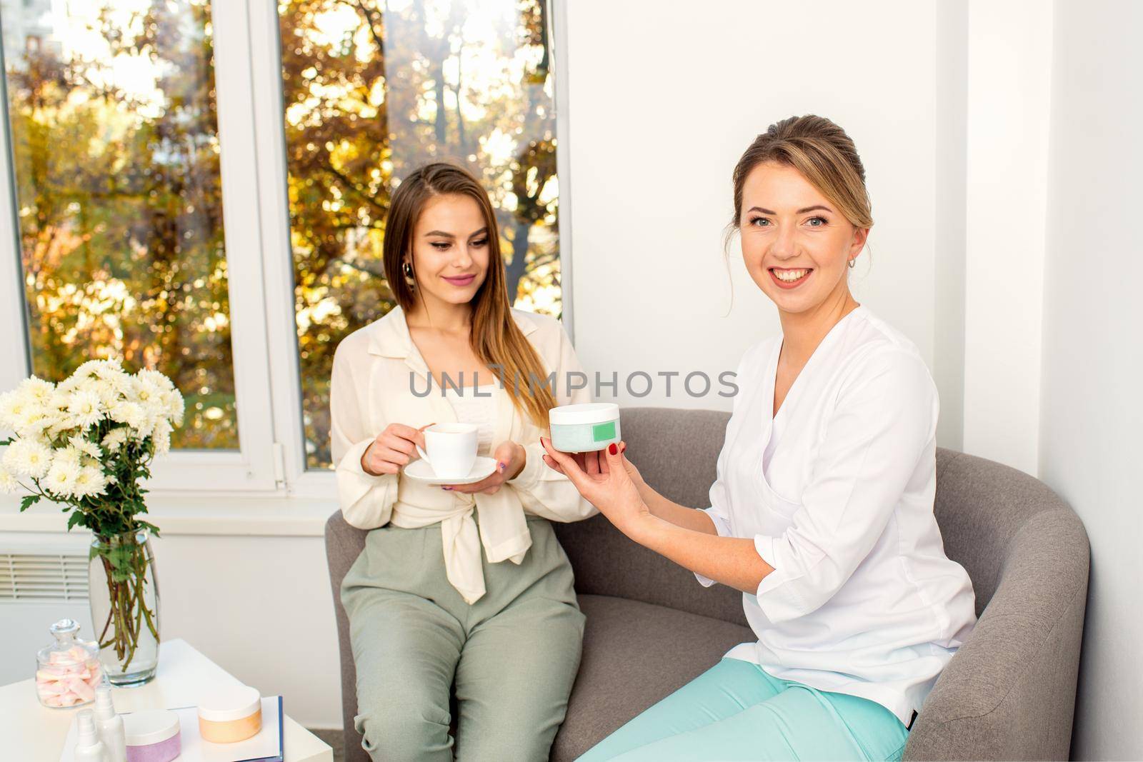 Beautician offering product for the young woman holding a white plastic jar with a cream sitting on the sofa