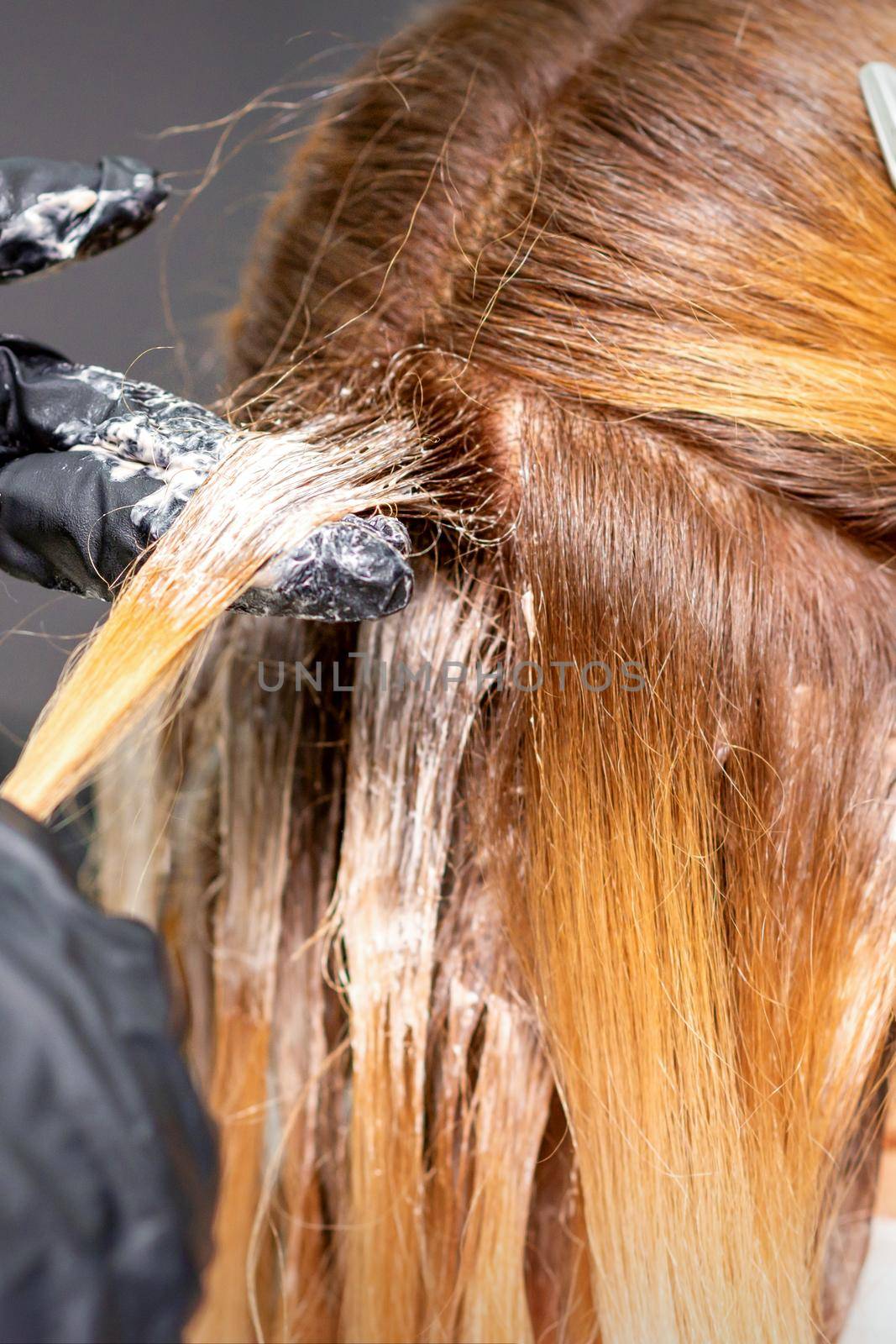 Hands of the hairdresser are applying the dye with gloved fingers on the red hair of a young red-haired woman in a hairdresser salon, close up. by okskukuruza