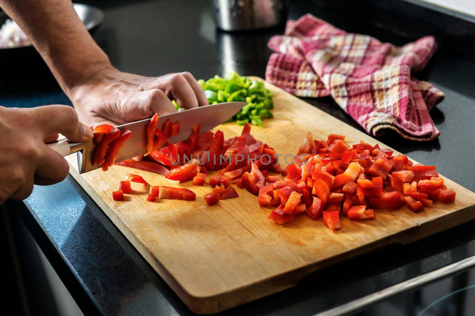 chef's hands chopping red pepper on a wooden cutting board, in the background you see chopped green pepper and a kitchen rag