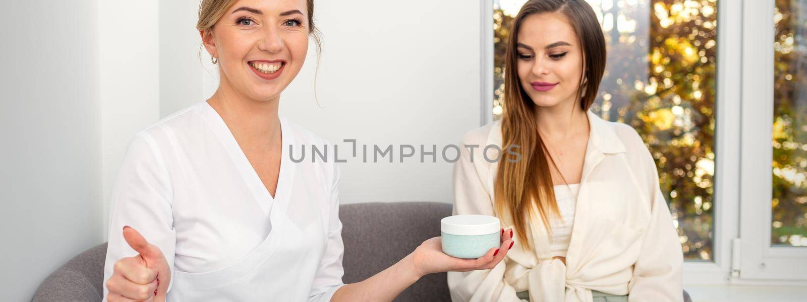 Beautician offering product for young woman, shows thumb up, holding a white plastic jar with a cream, mockup, copy space
