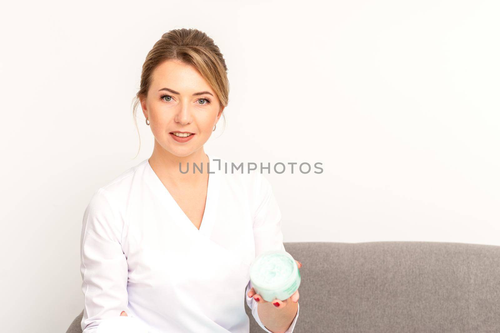 Close-up portrait of young smiling female caucasian healthcare worker standing with jar of cream beauty product on white background by okskukuruza