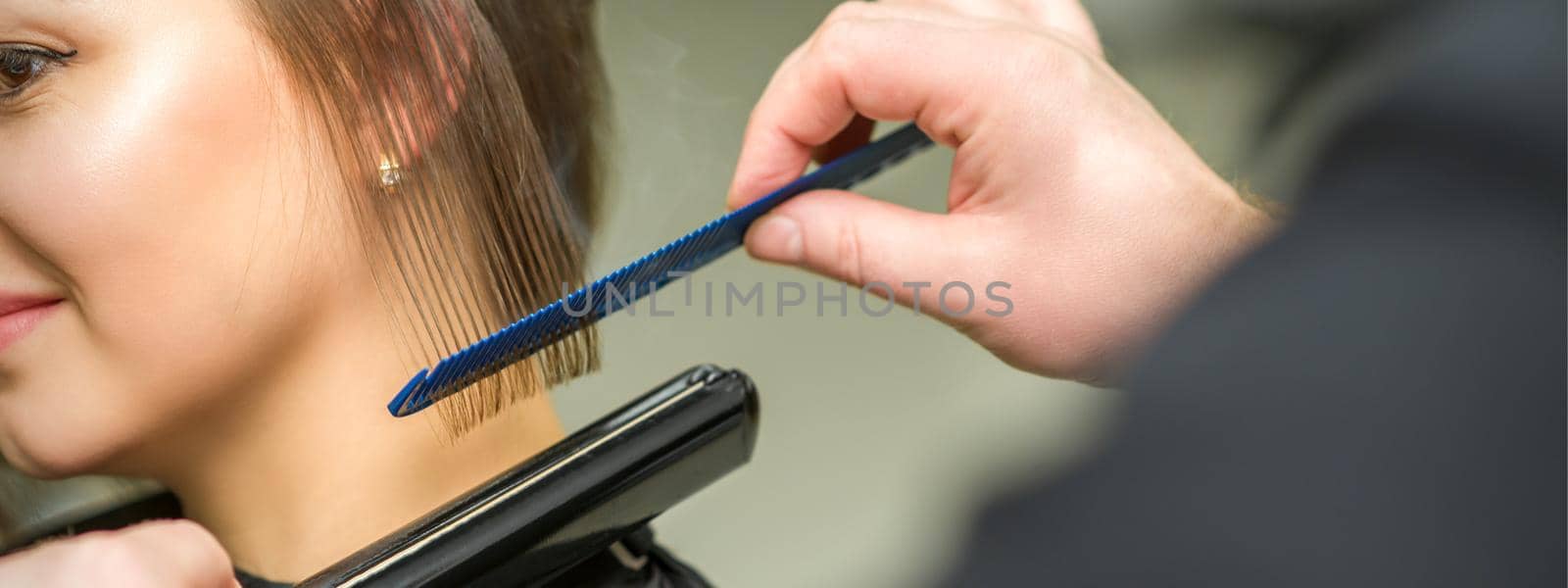 Hairstylist is straightening short hair of young brunette woman with a flat iron in a hairdresser salon, close up. by okskukuruza