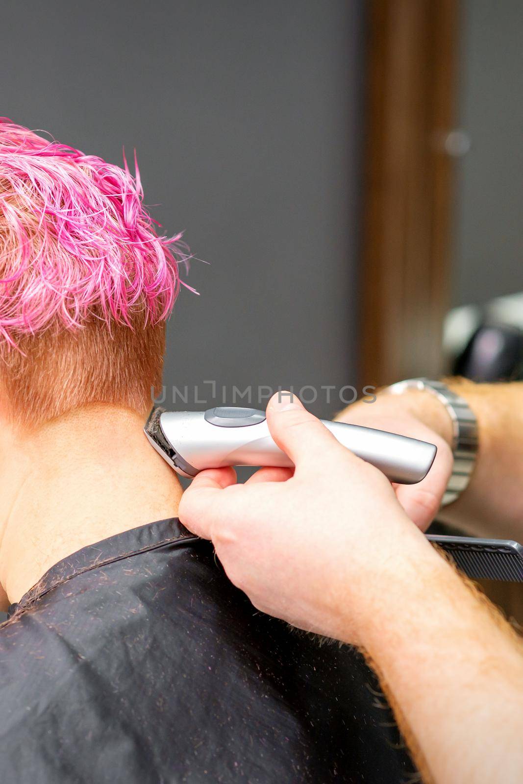 Male hairdresser shaves neck of a young caucasian woman with a short pink hairstyle by electric shaver in a hairdresser salon, close up